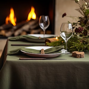 Pode incluir: Uma toalha de mesa de linho verde preparada para um jantar com pratos brancos, guardanapos verdes e um descanso de copo de madeira. A mesa est&aacute; posta em frente a uma lareira com fogo.