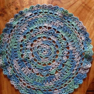 May include: A crocheted doily in shades of blue, green, and gray. The doily has a circular shape with a scalloped edge.
