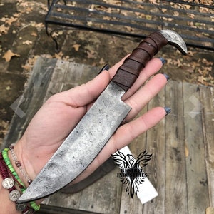 Pu&ograve; includere: Una mano tiene un coltello fatto a mano con un manico in pelle marrone e un pomo a forma di uccello. La lama &egrave; in acciaio e ha una finitura martellata. Il coltello &egrave; etichettato "Kestrel Industries".