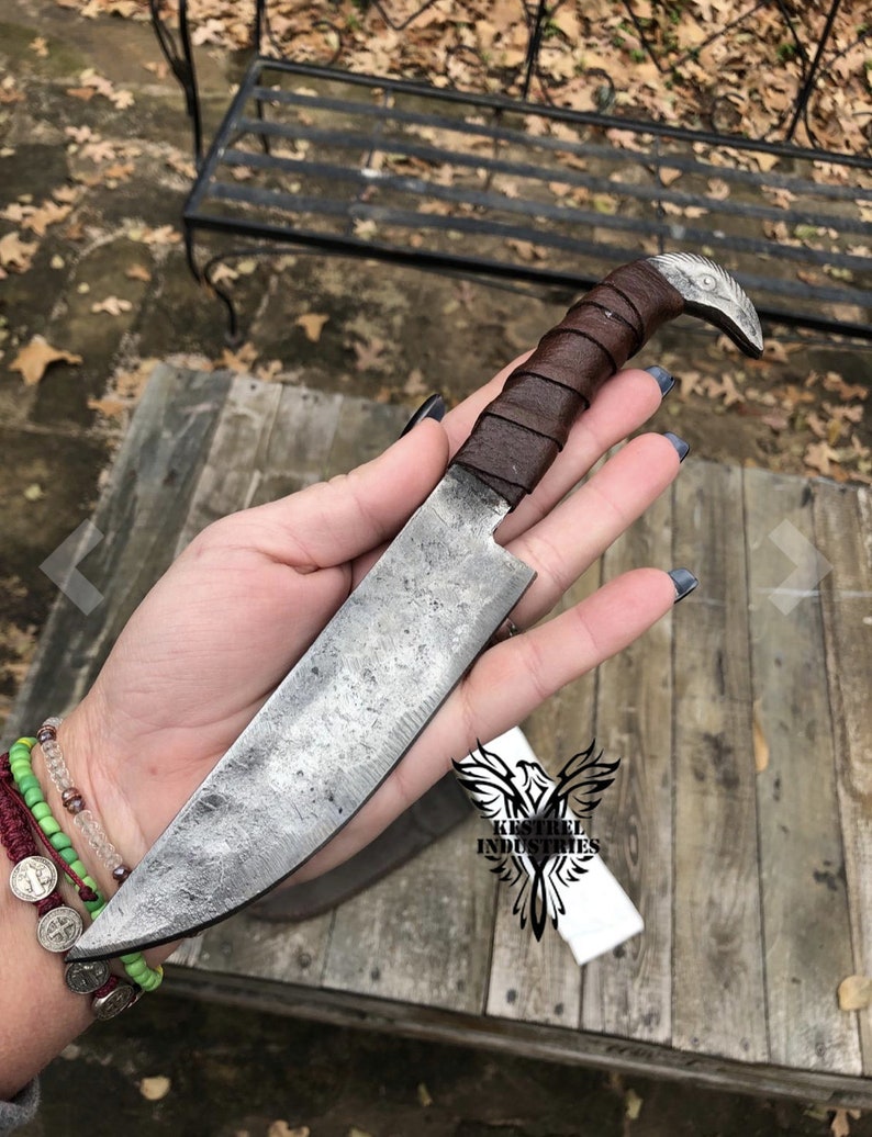 Pu&ograve; includere: Una mano tiene un coltello fatto a mano con un manico in pelle marrone e un pomo a forma di uccello. La lama &egrave; in acciaio e ha una finitura martellata. Il coltello &egrave; etichettato "Kestrel Industries".