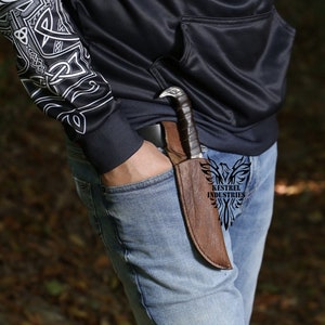 Pu&ograve; includere: Un fodero in pelle marrone con un coltello al suo interno. Il fodero &egrave; indossato su una cintura. Il coltello ha un'impugnatura a forma di uccello e una lama nera. Il testo "Kestrel Industries" &egrave; visibile sul fodero.