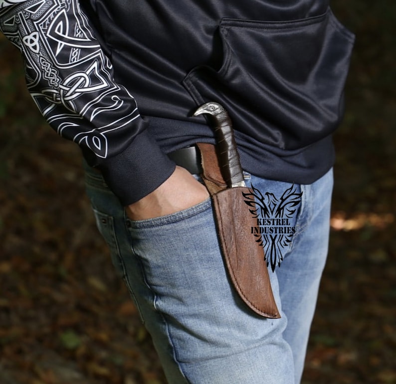 Pu&ograve; includere: Un fodero in pelle marrone con un coltello al suo interno. Il fodero &egrave; indossato su una cintura. Il coltello ha un'impugnatura a forma di uccello e una lama nera. Il testo "Kestrel Industries" &egrave; visibile sul fodero.