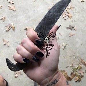 Pu&ograve; includere: Una mano che tiene un coltello forgiato nero con un manico in legno. Il coltello ha una lama curva e un gancio all'estremit&agrave; del manico. La mano ha lo smalto nero e un tatuaggio che dice "IV.VI". Il coltello ha un logo che dice "Kestrel Industries".