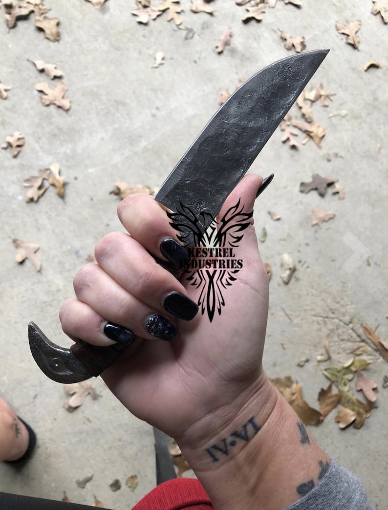 Pu&ograve; includere: Una mano che tiene un coltello forgiato nero con un manico in legno. Il coltello ha una lama curva e un gancio all'estremit&agrave; del manico. La mano ha lo smalto nero e un tatuaggio che dice "IV.VI". Il coltello ha un logo che dice "Kestrel Industries".