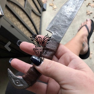 Pu&ograve; includere: Una mano che tiene un piccolo coltello fatto a mano con un manico in pelle marrone. La lama &egrave; in acciaio e ha una finitura martellata. Il coltello ha un manico unico a forma di uccello.