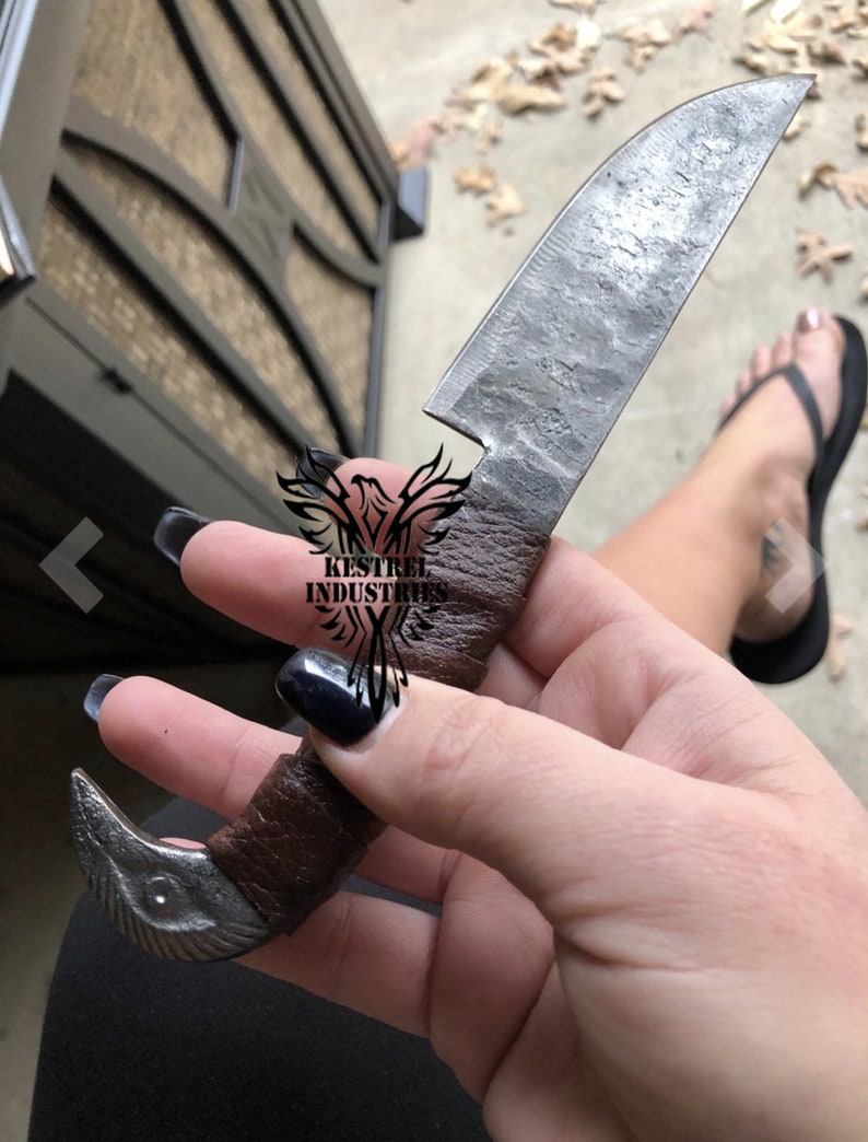 Pu&ograve; includere: Una mano che tiene un piccolo coltello fatto a mano con un manico in pelle marrone. La lama &egrave; in acciaio e ha una finitura martellata. Il coltello ha un manico unico a forma di uccello.