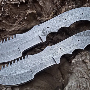 Può includere: Due coltelli in acciaio Damasco con bordi seghettati e lame decorate. I manici hanno scanalature per le dita. Le lame sono lunghe circa 25 cm. I coltelli sono esposti su una superficie di legno stagionato.