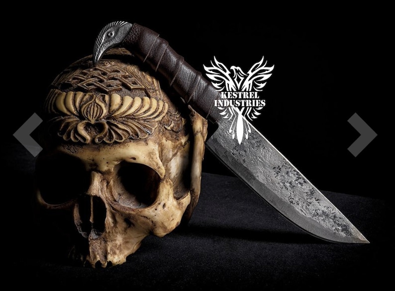Pu&ograve; includere: Un coltello forgiato a mano con un manico in pelle marrone e un teschio con intagli intricati. Il coltello &egrave; appoggiato sulla testa del teschio. La lama &egrave; in acciaio con una finitura martellata. Il logo "Kestrel Industries" &egrave; visibile sullo sfondo.