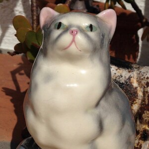 Puede incluir: Figura de gato de cerámica en posición sentada. El gato tiene un pelaje gris claro y blanco con orejas y nariz rosas. Los ojos son verdes. La figura es brillante y parece vintage.