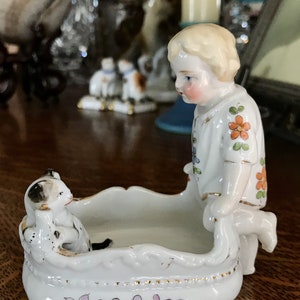 May include: A porcelain figurine of a child in a floral dress standing on a white porcelain dish with a black and white dog figurine inside. The dish has a floral design around the edge.