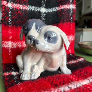 Puede incluir: Una figura de cerámica de dos cachorros, uno con una mancha gris sobre el ojo y la cabeza, el otro con una mancha gris. Los cachorros están sentados juntos, con un acabado brillante. La figura está sobre una tela a cuadros roja, negra y blanca.