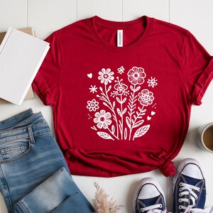 Könnte beinhalten: Rotes T-Shirt mit einem weißen Blumenmuster. Das Design zeigt stilisierte Blumen, Blätter und Herzen.