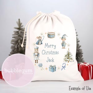 May include: A cream-coloured Christmas gift bag with a drawstring closure. The bag features a festive design with a circle of illustrations including a mouse, elf, nutcracker, and Christmas tree. The text "Merry Christmas Jack" is in the centre.
