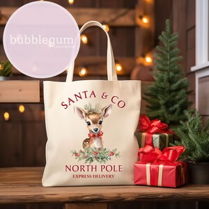 K&ouml;nnte beinhalten: Eine beige Canvas-Tragetasche mit einem rot-wei&szlig;en Design. Die Tasche zeigt ein Cartoon-Reh mit einer roten Schleife und den Text "Santa & Co. North Pole Express Delivery".