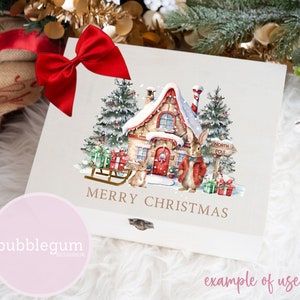 May include: A white wooden box with a red bow on top. The box has a watercolor illustration of a snow-covered cottage with a red door, two bunnies, a sled, and a sign that says "North Pole". The text "Merry Christmas" is written in gold lettering below the illustration.