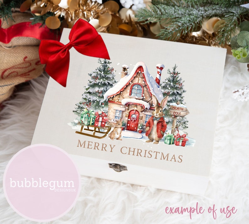 May include: A white wooden box with a red bow on top. The box has a watercolor illustration of a snow-covered cottage with a red door, two bunnies, a sled, and a sign that says "North Pole". The text "Merry Christmas" is written in gold lettering below the illustration.