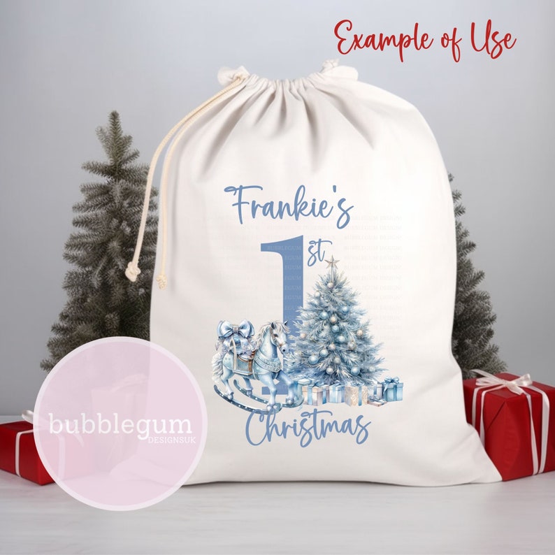 K&ouml;nnte beinhalten: Ein wei&szlig;er Stoff-Weihnachtssack mit blauem Text, der "Frankie's 1st Christmas" lautet. Der Sack zeigt ein blaues Schaukelpferd mit einer blauen Schleife und einen blauen Weihnachtsbaum mit blauen Kugeln und Geschenken.