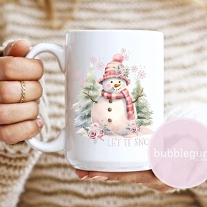 Puede incluir: Taza de cer&aacute;mica blanca con una ilustraci&oacute;n de acuarela de un mu&ntilde;eco de nieve con sombrero y bufanda rosas y rojos. La taza tiene el texto "LET IT SNOW" y est&aacute; decorada con rosas rosas y copos de nieve. La taza es sostenida por una persona.