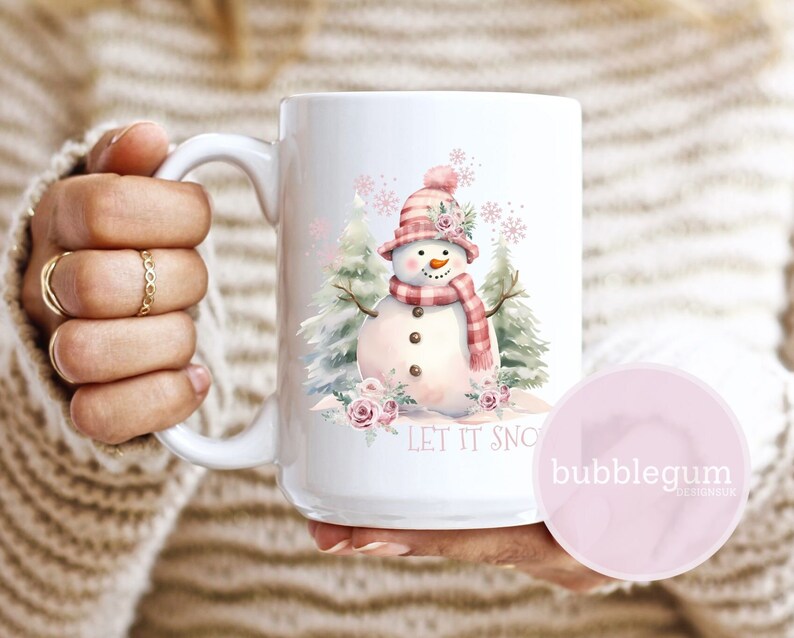Puede incluir: Taza de cer&aacute;mica blanca con una ilustraci&oacute;n de acuarela de un mu&ntilde;eco de nieve con sombrero y bufanda rosas y rojos. La taza tiene el texto "LET IT SNOW" y est&aacute; decorada con rosas rosas y copos de nieve. La taza es sostenida por una persona.