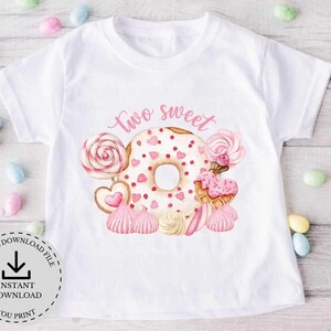 以下が含まれることがあります： 白いTシャツに、ピンクのドーナツと「two sweet」の文字がプリントされています。ドーナツはピンクと白のキャンディーとハートで囲まれています。