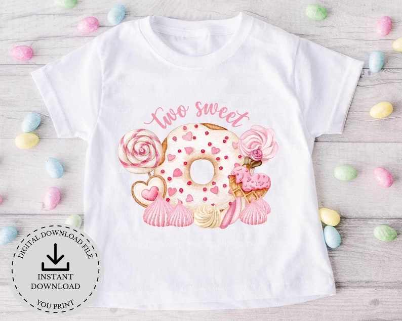 以下が含まれることがあります： 白いTシャツに、ピンクのドーナツと「two sweet」の文字がプリントされています。ドーナツはピンクと白のキャンディーとハートで囲まれています。