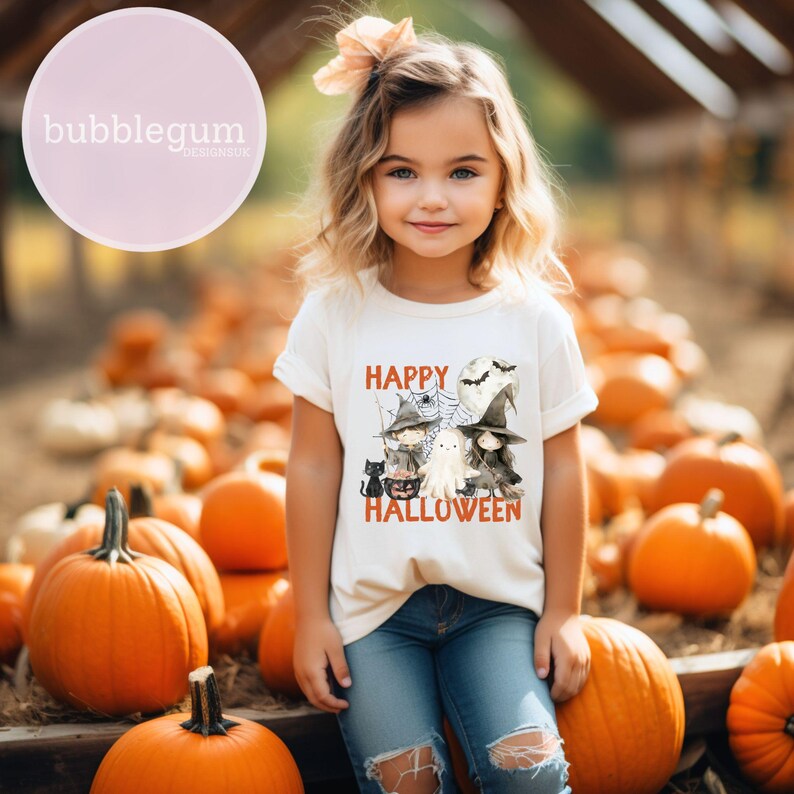 May include: A white t-shirt featuring a graphic of a group of cartoon witches, ghosts, and cats. The text "Happy Halloween" is printed on the shirt. A young girl is wearing the shirt and sitting amongst a pile of pumpkins.