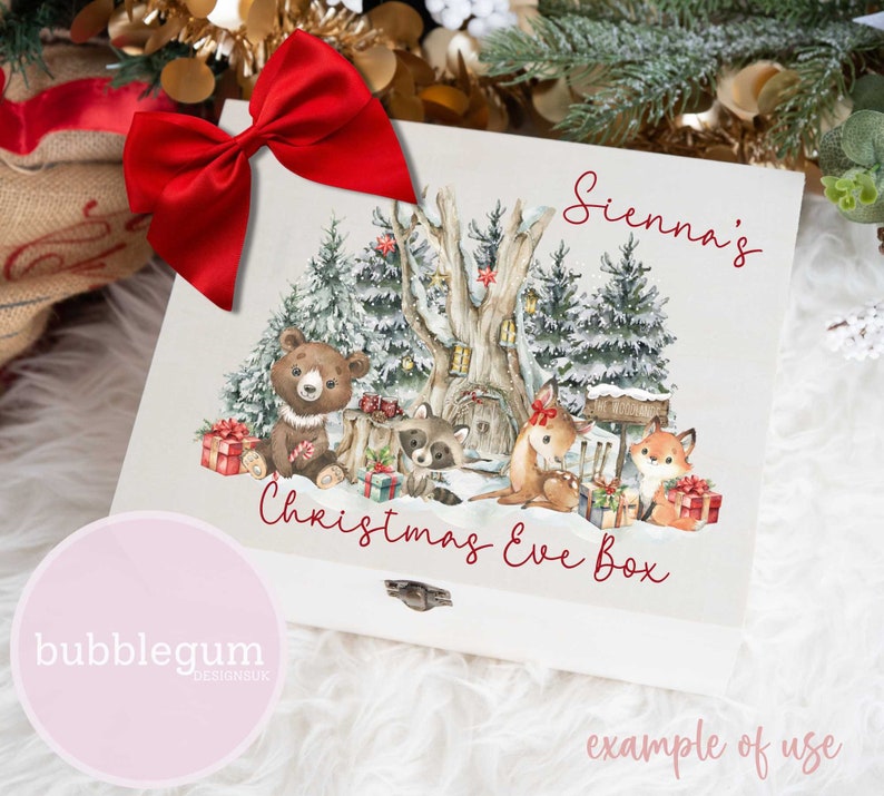 May include: A white wooden box with a watercolour illustration of a snowy forest scene with animals. The box has a red bow and the text "Sienna's Christmas Eve Box" in red lettering. The text "bubblegum designsuk" is in pink lettering on a pink circle.