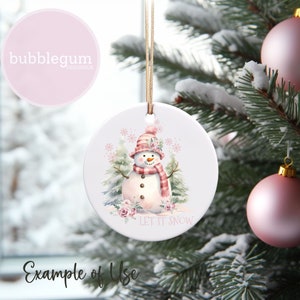 Puede incluir: Adorno navide&ntilde;o redondo y blanco con un mu&ntilde;eco de nieve de acuarela que lleva una bufanda y un gorro de cuadros rosas y rojos, con el texto "LET IT SNOW". El adorno cuelga de un hilo dorado, con una bola rosa en el fondo.