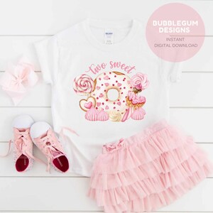 以下が含まれることがあります： 白いTシャツにピンクのドーナツと「two sweet」の文字が書かれています。ドーナツはピンクのハートとスプリンクルで飾られています。ドーナツの周りには、ロリポップ、ハート型のクッキー、メレンゲなど、他のピンクのスイーツがあります。
