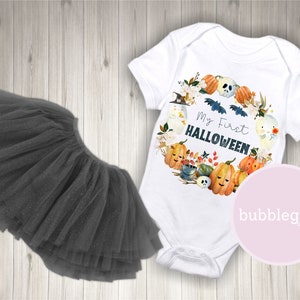 Pode incluir: Um macac&atilde;o branco para beb&ecirc; com um design de aquarela de ab&oacute;boras, fantasmas e morcegos. O macac&atilde;o diz "Meu primeiro Halloween". Tamb&eacute;m &eacute; mostrado um tutu de tule preto.