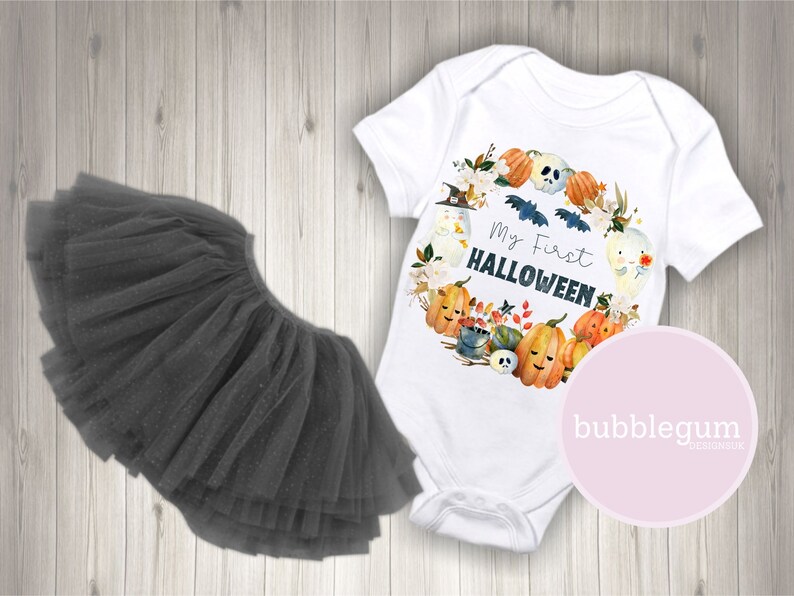 Pode incluir: Um macac&atilde;o branco para beb&ecirc; com um design de aquarela de ab&oacute;boras, fantasmas e morcegos. O macac&atilde;o diz "Meu primeiro Halloween". Tamb&eacute;m &eacute; mostrado um tutu de tule preto.