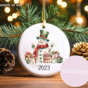 May include: White ceramic Christmas ornament with a watercolor illustration of a snowman wearing a red scarf and green hat, surrounded by presents and candy canes. The ornament has the year 2023 printed on it.