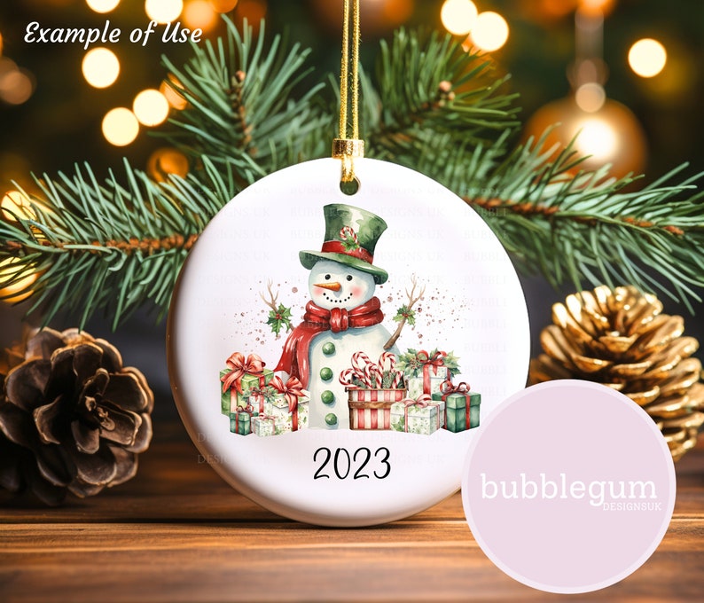 May include: White ceramic Christmas ornament with a watercolor illustration of a snowman wearing a red scarf and green hat, surrounded by presents and candy canes. The ornament has the year 2023 printed on it.