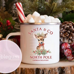 K&ouml;nnte beinhalten: Wei&szlig;e Emaille-Tasse mit einer rot-gr&uuml;nen Illustration eines Rehs mit einer roten Schleife. Die Tasse tr&auml;gt den Schriftzug "Santa & Co. Nordpol Expresslieferung".