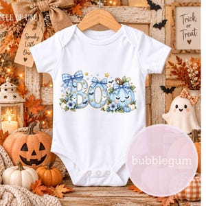 May include: A white short-sleeved baby bodysuit featuring a graphic print of the word BOO in light blue with a plaid pattern, decorated with blue bows, small ghosts, bats, stars, and a light blue pumpkin with a smiling face and floral accents.