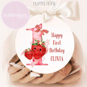 May include: A clear cellophane bag filled with cookies, tied with a cream ribbon. A white circular tag features a pink number one, strawberries, and the text "Happy First Birthday OLIVIA". The tag also has the text "bubblegum DESIGNSUK".