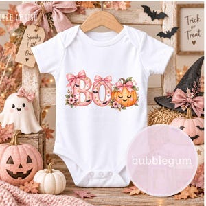 May include: A white short-sleeved baby bodysuit featuring a graphic print of the word BOO in pink letters decorated with bows, flowers, and small bats, with an orange pumpkin replacing the letter O, all centered on the front of the garment.