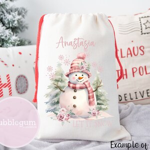 Puede incluir: Bolsa de regalo con cord&oacute;n de color crema con un mu&ntilde;eco de nieve de acuarela con sombrero y bufanda rosas. La bolsa tiene el nombre "Anastasia" y la frase "Let It Snow" impresa. Cierre con cord&oacute;n rojo.