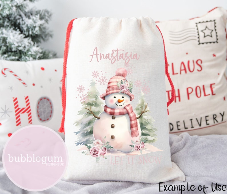 Puede incluir: Bolsa de regalo con cord&oacute;n de color crema con un mu&ntilde;eco de nieve de acuarela con sombrero y bufanda rosas. La bolsa tiene el nombre "Anastasia" y la frase "Let It Snow" impresa. Cierre con cord&oacute;n rojo.