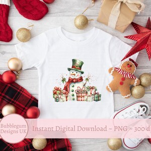 May include: A white t-shirt with a watercolor illustration of a snowman holding candy canes and surrounded by wrapped presents. The snowman is wearing a green hat and a red scarf. The background is a white wooden surface with Christmas decorations.