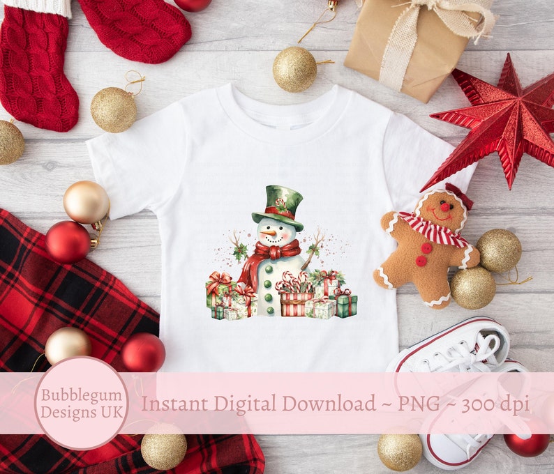 May include: A white t-shirt with a watercolor illustration of a snowman holding candy canes and surrounded by wrapped presents. The snowman is wearing a green hat and a red scarf. The background is a white wooden surface with Christmas decorations.