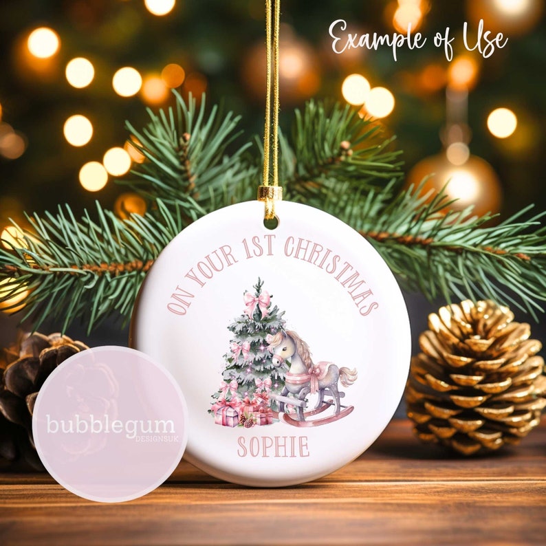 May include: White ceramic Christmas ornament with a pink and green illustration of a Christmas tree, a rocking horse, and presents. The text "On Your 1st Christmas Sophie" is written in pink around the illustration.