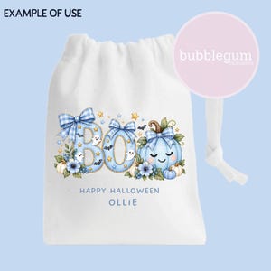May include: A white drawstring bag featuring a blue graphic with the text BOO, HAPPY HALLOWEEN, and OLLIE. The design includes a light blue pumpkin with a face, blue plaid bows, small ghosts, bats, stars, and floral accents in shades of blue.