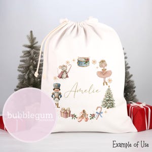 May include: Cream-colored drawstring gift bag featuring a watercolor design of ballet-themed elements. The design includes a ballerina, nutcracker, mouse, drum, and Christmas tree. The name "Amelie" is written in gold script. The bag has a white drawstring closure.