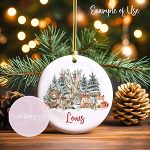 May include: A white ceramic Christmas bauble featuring a winter woodland scene with a bear, raccoon, fox, and deer. The bauble is personalised with the name "Louis".