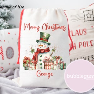 May include: A white canvas drawstring bag with a red drawstring. The bag features a watercolor illustration of a snowman with a green hat, red scarf, and candy canes. The text "Merry Christmas George" is printed on the bag.