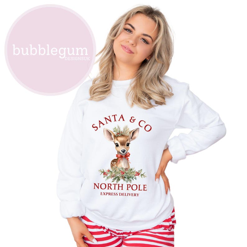 K&ouml;nnte beinhalten: Wei&szlig;er Weihnachtspullover mit einer rot-gr&uuml;nen Illustration eines Rehs mit einer roten Schleife und dem Text "Santa & Co. North Pole Express Delivery".