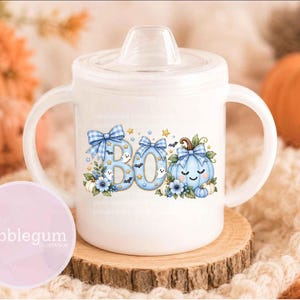 May include: A white sippy cup with two handles and a clear lid, featuring a graphic of the word BOO in light blue plaid letters, decorated with small ghosts, bats, stars, blue flowers, and a light blue plaid pumpkin with a smiling face.