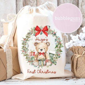 May include: A cream-colored drawstring gift bag with a Christmas-themed design. The design includes a teddy bear, a red bow, and the text "Hugo's First Christmas". The bag is decorated with a wreath of greenery and berries.