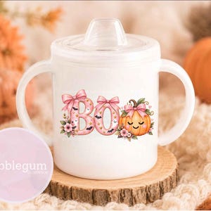 May include: A white sippy cup with two handles and a clear lid, featuring a pink graphic that spells BOO with bows, small ghosts, bats, and a smiling pumpkin with a pink bow, decorated with floral accents and gold stars on the front surface.